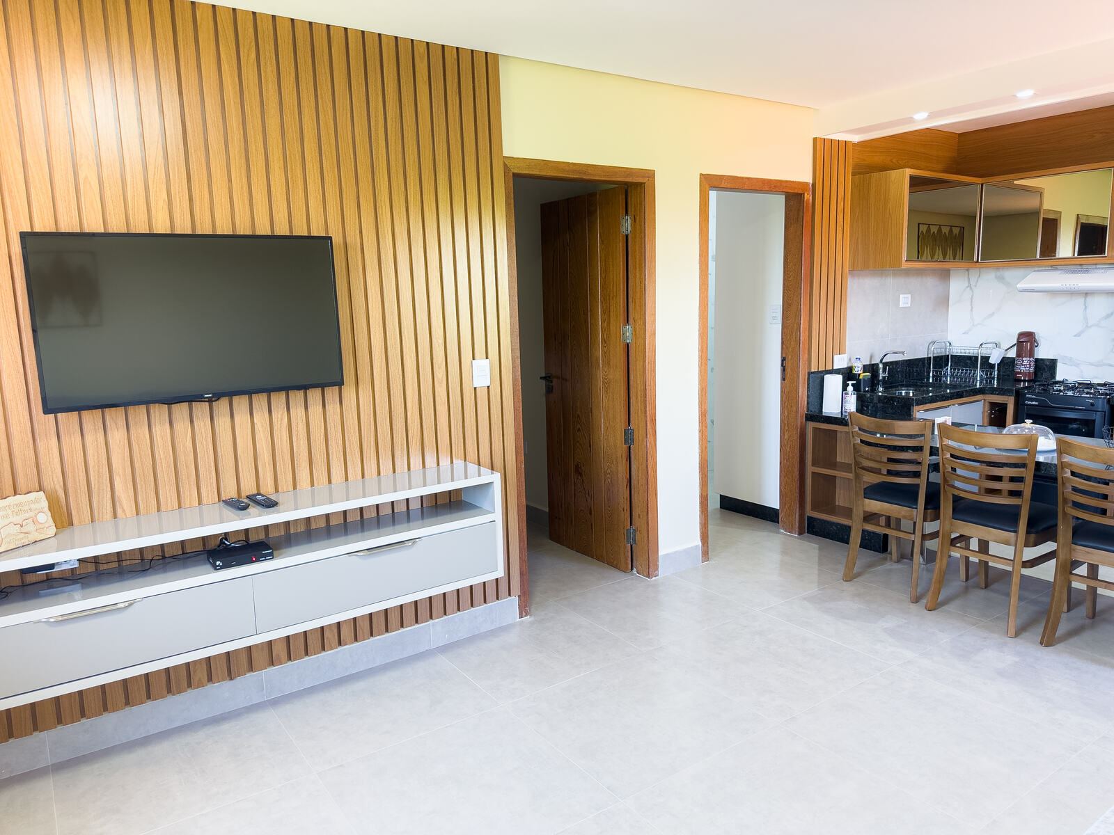Sala com TV em painel ripado de madeira e, ao fundo, a cozinha e a mesa de jantar